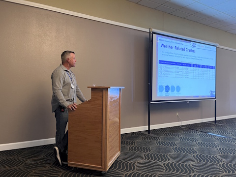A man speaks standing behind a podium, slide with 'Weather Related Crashes' displayed on a projector screen on the right.